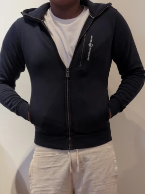 Sail racing  - Snygg marinblå hoodie från Sail Racing med dragkedja framtill och huva. Diskret logga på bröstet och ribbade muddar vid ärmar och midja. Tillverkad i mjukt material som känns skönt mot huden. Perfekt för en avslappnad och sportig stil.