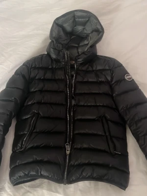Colmar jacka svart  - Säljer en svart pufferjacka med huva och två sidofickor med dragkedja. Jackan har en glansig finish, quiltad design och en patch på ärmen. Perfekt för kalla dagar och riktigt snygg till streetwear-stilen.