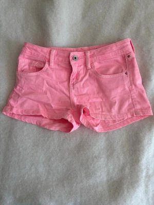 Rosa shorts från Guess - Snygga rosa shorts från Guess i klassisk femficksmodell. De har bälteshällor, knapp och dragkedja framtill samt den ikoniska Guess-loggan på bakfickan. Perfekta för varma dagar och ger en fräsch look till din outfit.