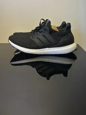 Svarta Adidas Ultraboost sneakers - Säljer ett par svarta Adidas Ultraboost sneakers med stickad ovandel och vita Boost-sulor. Skorna har klassiska tre ränder på sidan, svart hälkappa och slitstark yttersula med grepp. Perfekta för dig som gillar stilrena och bekväma sneakers.