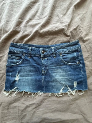 Blå jeanskjol med fransig kant - Säljer en blå jeanskjol i kort modell med slitna detaljer och fransig nederkant. Kjolen har klassiska fickor fram och bak samt bälteshällor. Perfekt för dig som gillar en avslappnad och trendig stil.