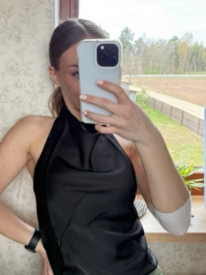 Svart halterneck topp med snörning - Snygg svart halterneck topp i satinliknande material med snörning i ryggen. Toppen är ärmlös och har en croppad passform som ger en trendig look. Perfekt att styla med jeans eller kjol för en cool outfit.
