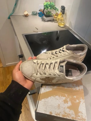 Vita sneakers med glitterstjärna - Säljer ett par vita sneakers med en glittrig guldstjärna på sidan och vita snören. Skorna har en klassisk låg profil och detaljer i mocka på tån. Märket syns på plösen och designen är trendig och ungdomlig.
