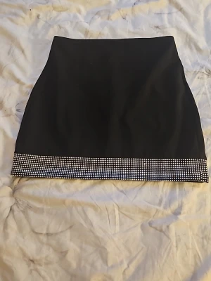 Svart minikjol med glittrig kant - Snygg svart minikjol med rak passform och glittrig kant nertill i silverfärgade stenar. Kjolen har en enkel design som gör den lätt att matcha med olika toppar. Materialet ser ut att vara ett mjukt tyg med lite stretch.