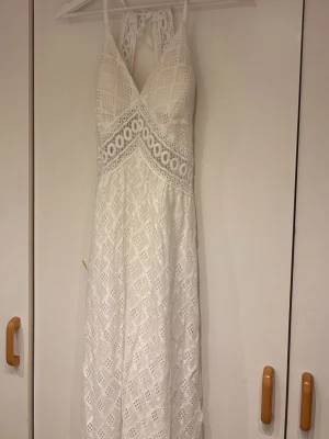 Vit lång spetsklänning med halterneck - Säljer en elegant vit långklänning med spetsdetaljer och halterneck. Klänningen har v-ringad urringning, öppen rygg och fina broderade mönster. Perfekt för sommarens alla tillfällen och studentfirande.
