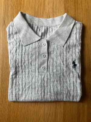 Ljusgrå kabelstickad polotopp - Säljer en ljusgrå kabelstickad polotopp med korta ärmar och klassisk krage. Toppen har tre knappar framtill och en diskret broderad logga på bröstet. Perfekt för dig som gillar stilrena och tidlösa plagg.