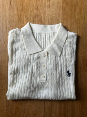 Vit kabelstickad polotopp från Ralph Lauren - Vit kabelstickad polotopp från Ralph Lauren med korta ärmar och klassisk krage. Toppen har tre knappar framtill och den ikoniska mörkblå loggan broderad på bröstet. Perfekt till jeans eller kjol för en clean och stilren look.