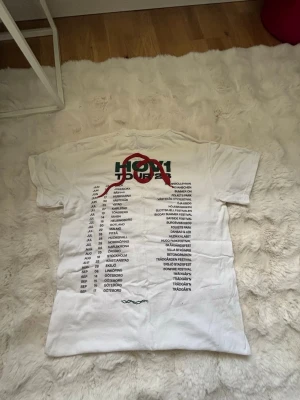 Vit t-shirt med tryck Hov1 Tour - Vit t-shirt med Hov1 Tour-tryck på ryggen och liten krona på bröstet. Klassisk passform med rund hals och korta ärmar.  Har några små fläckar därför jag säljer billigare går även att diskutera priset. 