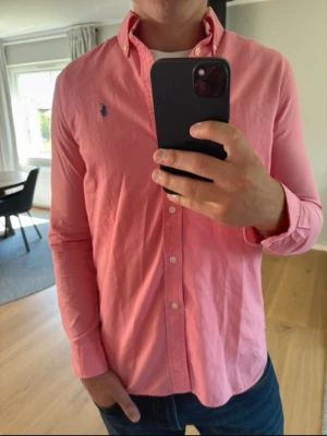 Rosa skjorta med knappkrage - Säljer en stilren rosa skjorta med knappkrage och broderad logga på bröstet. Skjortan har långa ärmar, klassisk passform och knäppning framtill. Perfekt för dig som vill ha en fräsch och enkel look.
