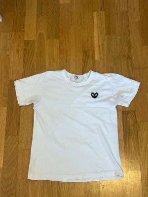 Vit t-shirt med svart hjärtdetalj - Vit t-shirt med rund hals och korta ärmar från  Comme des Garçons. På bröstet sitter en svart hjärtdetalj med ögon, vilket ger en unik och trendig look. Tillverkad i mjuk bomull som känns skön mot huden. Perfekt för dig som gillar minimalistisk stil med en twist. Den är i storlek L men skulle passa 164-170 cm. Skriv till mig om du har några frågor. 