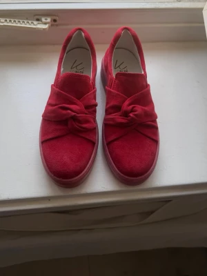 Röda slip-on skor med knut - Snygga röda slip-on skor i mockaimitation med en unik knutdetalj på ovansidan. Skorna har en tjock sula i matchande röd färg och en stilren, modern look. Perfekta för dig som vill sticka ut med dina skor och gillar trendiga detaljer.