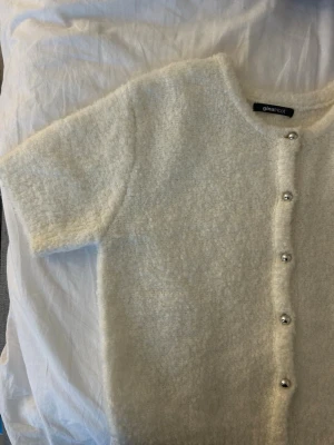Vit fluffig kofta från Gina Tricot - Kortärmad, vit kofta från Gina Tricot i fluffigt material. Koftan har rund halsringning och knäpps med silvriga knappar framtill. Perfekt för lager på lager och ger en mysig look till din outfit.