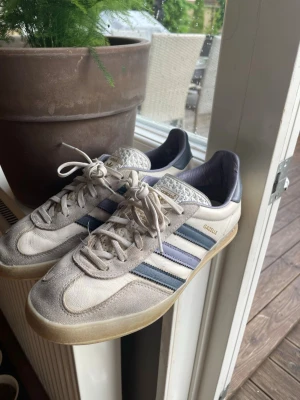Adidas Gazelle Indoor White/Blå - Säljer ett par Adidas Gazelle Indoor sneakers i vitt och blått med klassiska tre ränder på sidan. Ovandelen är i mocka och läder, med gummisula och guldfärgad Gazelle-text på sidan. De har varit inne hos en skomakare, ganska sällsynta, st 41 1/3.