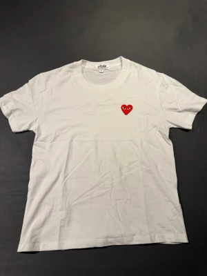 Vit t-shirt Comme des Garçons Play - Vit t-shirt från Comme des Garçons Play med det ikoniska röda hjärtat broderat på bröstet. Klassisk passform och rund halsringning, tillverkad i mjuk bomull. Perfekt för en clean och stilren look.