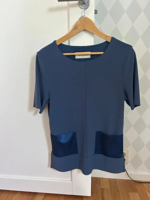 Blus By Malene Birger - Stilren blå blus med korta ärmar och rund halsringning. Två snygga fickor framtill i blankt satinmaterial ger en cool kontrast, dragkedja på båda sidorna! Storleken är xxs men passar dig som vanligtvis är xs/s. 