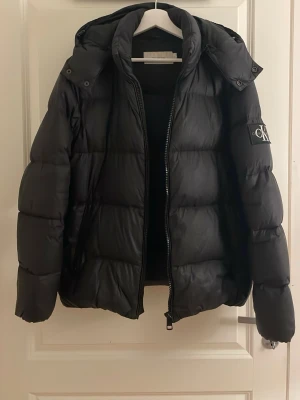 Svart pufferjacka från Calvin Klein - Snygg svart pufferjacka från Calvin Klein med huva och dragkedja framtill. Jackan har en patch med CK-logga på ärmen och breda, vadderade paneler som ger en riktigt puffig look. Perfekt för kalla vinterdagar och enkel att matcha med olika outfits.