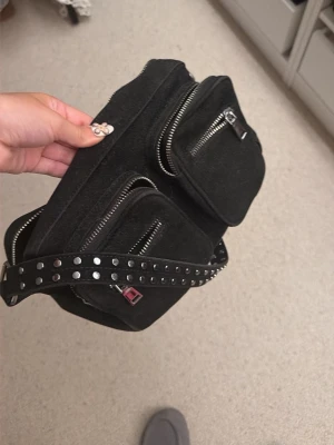 Svart väska med nitar - Säljer en svart väska i tyg med två framfickor och silvriga dragkedjor. Väskan har ett justerbart band dekorerat med silverfärgade nitar för en cool look. Perfekt för att bära det viktigaste och ger en edgy vibe till outfiten.