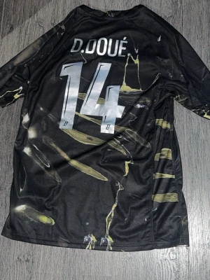 Svart PSG matchtröja D. Doué 14 - Säljer en svart PSG matchtröja med namn och nummer D. Doue 14 på ryggen. Tröjan har ett unikt mönster i gult och vitt, samt Qatar Airways-logga på bröstet. Tillverkad i lätt och ventilerande material, perfekt för fotboll eller träning.