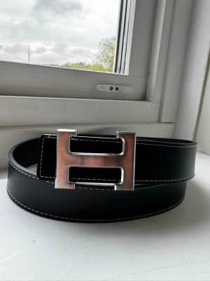 Svart skärp med silverfärgat H-spänne - ❗️FAST PRIS❗️Stilrent svart bälte med ett stort, silverfärgat H-format spänne. Bältet har vit kontrastsöm längs kanterna och är tillverkat i skinn med en lätt texturerad yta. Perfekt accessoar för att lyfta din outfit.