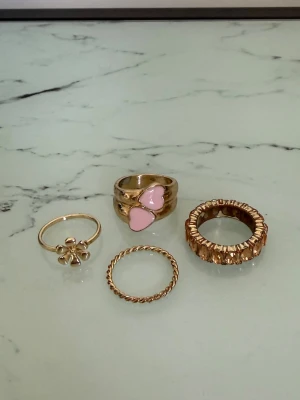 Fyra guldfärgade ringar med detaljer - Säljer ett set med fyra guldfärgade ringar. En ring har två rosa hjärtan, en har en liten blomma, en är tvinnad och en har en bred, strukturerad design. Perfekt för dig som gillar att mixa och matcha olika stilar. Ringarna är i storlek L och är inte i äkta guld, så de har börjat rosta. 