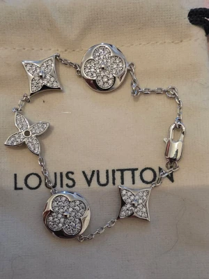 Louis Vuitton armband med kristaller - Snyggt armband från Louis Vuitton i silverfärg med flera ikoniska blommor och stjärnor dekorerade med gnistrande kristaller. Länken är tunn och smidig, och detaljerna är riktigt blingiga. Perfekt accessoar för dig som gillar lyxig och trendig stil.