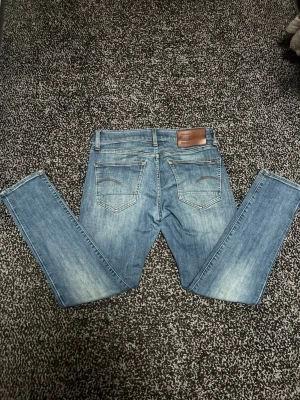 Blå jeans från G-Star RAW - Snygga blå jeans från G-Star RAW med klassisk femficksdesign och ljus tvätt. Jeansen har raka ben och detaljerade sömmar på bakfickorna samt en brun läderpatch med logga i midjan. Perfekta för en avslappnad och trendig look.
