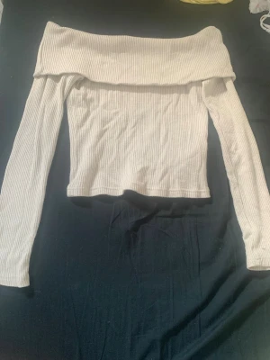 Vit offshoulder topp Gina Tricot - Trendig vit ribbad offshoulder topp från Gina Tricot. Toppen har långa ärmar och en bred kant över axlarna som ger en snygg och modern look. Perfekt att styla med både jeans och kjol för en cool outfit.