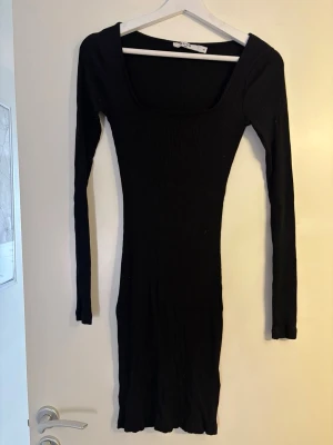 Svart ribbad långärmad klänning - Säljer en svart, ribbad klänning med lång ärm och fyrkantig urringning. Klänningen är figurnära och har en enkel, stilren design som passar perfekt till många olika tillfällen. Materialet känns mjukt och stretchigt.