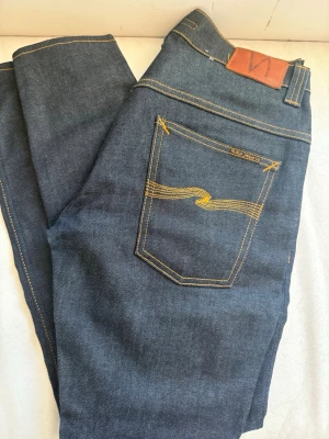 Mörkblå jeans från Nudie Jeans - Säljer ett par mörkblå jeans från Nudie Jeans med klassisk gul kontrastsöm och vågig brodyr på bakfickan. Jeansen har en lite mer slim passform och är tillverkade i kraftig denim. Perfekta för dig som gillar stilrena och tidlösa jeans. Tape Ted e modellen 