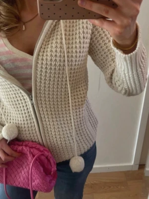 Vit stickad hoodie med dragkedja - Mysig vit stickad hoodie med dragkedja framtill och stora tofsar i snörena. Tröjan har en luva och ett grovt stickat mönster som ger en avslappnad look. Perfekt att slänga på sig över en t-shirt för extra värme.