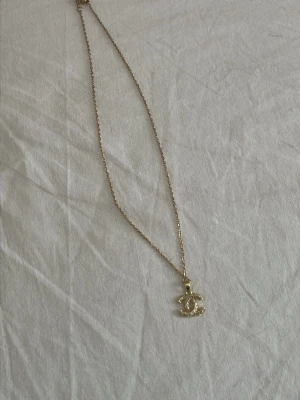 Guldigt halsband med glittrande hänge - Snyggt halsband i guldfärg med ett glittrande hänge i form av två sammanlänkade C:n, dekorerat med små pärlliknande stenar. Kedjan är tunn och elegant, perfekt för att lyfta vilken outfit som helst.