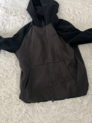 Svart och mörkgrå hoodie från Nike - Snygg hoodie från Nike i svart och mörkgrått med dragkedja framtill och två stora fickor. Tröjan har huva och är tillverkad i mjukt bomullsmaterial. Perfekt för en avslappnad streetwear-look.