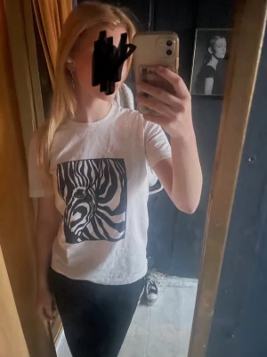 Vit t-shirt med zebramönster - Säljer en vit t-shirt med ett stort zebramönster i svart på bröstet. T-shirten har rund hals och korta ärmar. Perfekt för dig som gillar djurprint och vill ha en enkel men cool look.