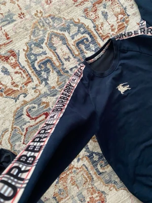 Mörkblå sweatshirt från Burberry - Snygg mörkblå sweatshirt från Burberry med klassisk logga broderad på bröstet och märkestext längs ärmarna. Tröjan har rund halsringning och långa ärmar med ribbade muddar. Perfekt för en trendig och avslappnad stil.