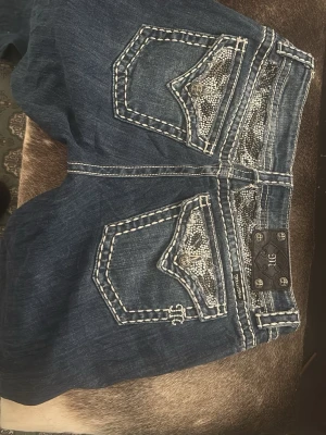 Mörkblå jeans med mönstrade fickor - Säljer ett par mörkblå jeans med vita kontrastsömmar och detaljerade bakfickor med ormmönstrat tyg. Jeansen har en klassisk femficksmodell och dekorativa knappar bak. Perfekta för dig som gillar unika detaljer och streetstyle.