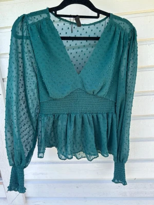 Grön blus med struktur från Amisu - Snygg grön blus från Amisu med v-ringning, transparenta långärmar och strukturprickar. Smockad midja och ärmslut ger en trendig siluett. Perfekt att styla med jeans eller kjol för en cool look.