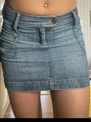 Blå jeanskjol med fickor - Snygg blå jeanskjol i kort modell med klassiska fickor fram och bak samt bälteshällor. Kjolen har knapp och dragkedja framtill och är tillverkad i denim. Perfekt för en trendig och avslappnad stil.