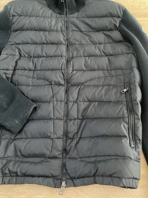 Moncler cardigan  - Säljer en svart cardigan från Moncler med quiltad front och stickade ärmar. Jackan har dragkedja framtill, hög krage och en ficka med dragkedja på sidan. Moncler-logga på ena ärmen ger en exklusiv touch. Perfekt för kallare dagar. Liten lagning på axeln och slutet av armen. Såklart äkta. Storlek Xl men sitter mer som en M/L