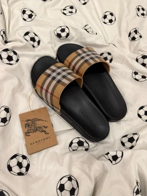Burberry sandaler med rutigt mönster - Snygga sandaler från Burberry med klassiskt beige, svart, vitt och rött rutmönster på remmen och svart sula. Perfekta för sommaren och riktigt trendiga. Tillverkade i syntetmaterial med präglad Burberry-logga på fotbädden.