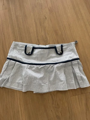 Randig kjol med marinblå detaljer - Säljer en vit kjol med tunna blå ränder och marinblå detaljer. Kjoldelen har veck och dekorativa knappar framtill samt snygga banddetaljer bak. Dragkedja i sidan och mjukt, lätt material. Från märket Pimkie