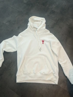 Vit hoodie med röd hjärtdetalj - Säljer en stilren vit hoodie med huva och dragsnören från Ami Paris. Hoodien har en klassisk känguruficka framtill och ribbade muddar vid ärmar och nederkant. 