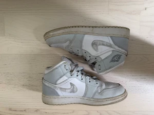 Nike Air Jordan 1 Mid vita/grå - Snygga Nike Air Jordan 1 Mid sneakers i vitt och ljusgrått med klassisk swoosh och Jordan Wings-logga på sidan. Skorna har en hög siluett och är tillverkade i läder och textil. Perfekta för dig som gillar streetwear och sportig stil. Kan tvättas innan dom skickas