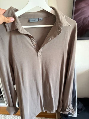 Beige henley från Snoot - Säljer en beige henley från Snoot med lång ärm och klassisk krage. Skjortan har knappar upptill och är gjord i mjukt bomullsmaterial. Perfekt för dig som gillar en enkel men stilren look.