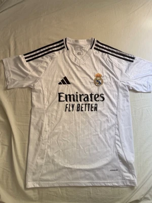 Real Madrid Hemma tröja 24/25 -  Real Madrid 24/25 Hemmatröja 🤍  Säljer en riktigt snygg Real Madrid 24/25 hemmatröja i storlek L passar även M. Tröjan är endast använd en gång, så den är i nästintill nyskick.  Säljer den eftersom jag helt enkelt glömt bort att jag hade den och den blir därför bara liggande. Perfekt för dig som vill ha en stilren fotbollströja eller supporta världens största klubb.