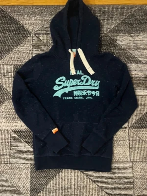 Mörkblå Superdry hoodie med tryck - Snygg mörkblå hoodie från Superdry med ljusblått tryck på bröstet och vita dragsnören i huvan. Klassisk känguruficka framtill och ribbade muddar. Perfekt för en avslappnad och cool stil.