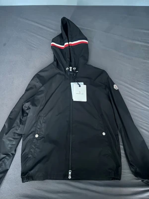 Svart vindjacka från Moncler - Snygg svart vindjacka från Moncler med huva och dragkedja framtill. Jackan har en röd och vit rand på huvan, två fickor med tryckknappar och Moncler-logga på ärmen. Tillverkad i lätt och smidigt material, perfekt för blåsiga dagar. Den är använd några gånger men de är knappt märkbart! 