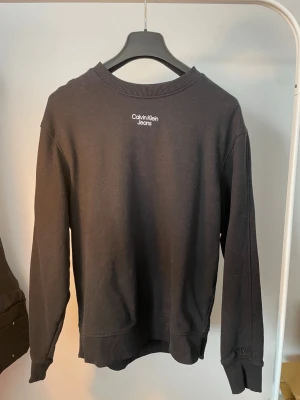 Svart sweatshirt från Calvin Klein Jeans - En svart sweatshirt från Calvin Klein Jeans med diskret logga broderad i vitt på bröstet. Tröjan har rund halsringning, ribbade muddar vid ärmslut och nederkant samt en klassisk, avslappnad passform. Perfekt till ett par jeans eller joggers.