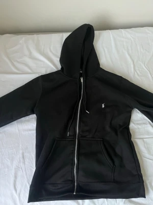 Svart zip hoodie med huva - Snygg svart hoodie med dragkedja och huva. Klassisk design med fickor framtill och justerbara snören i huvan. Liten vit logga broderad på bröstet. Perfekt för en chill och avslappnad stil. Tillverkad i mjukt material för extra komfort.