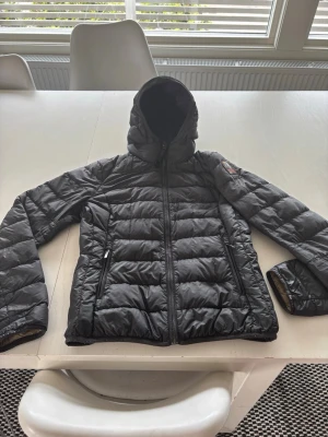 parajumper - Snygg svart dunjacka med huva och dragkedja framtill. Jackan har två sidofickor med dragkedja och quiltad design. Perfekt för kalla vinterdagar och har en patch på ärmen för extra stil.