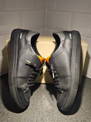 Svarta Axel Arigato sneakers - Snygga svarta sneakers från Axel Arigato med orange plös och svarta skosnören.